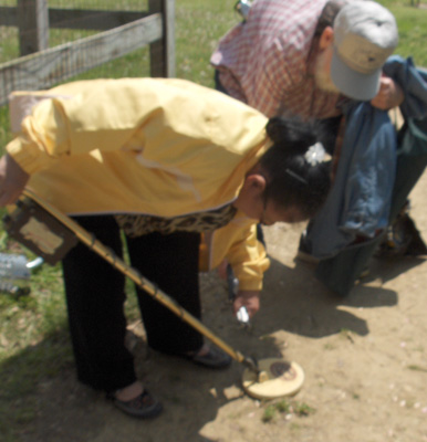 3 photos show Nanta and a man standing on a path.  Nanta is holding the metal detector (looks like a long stick with a round plate on the bottom).  The middle photo shows her bending down to reach under the plate, and the 3rd photo shows her picking up her folded cane.